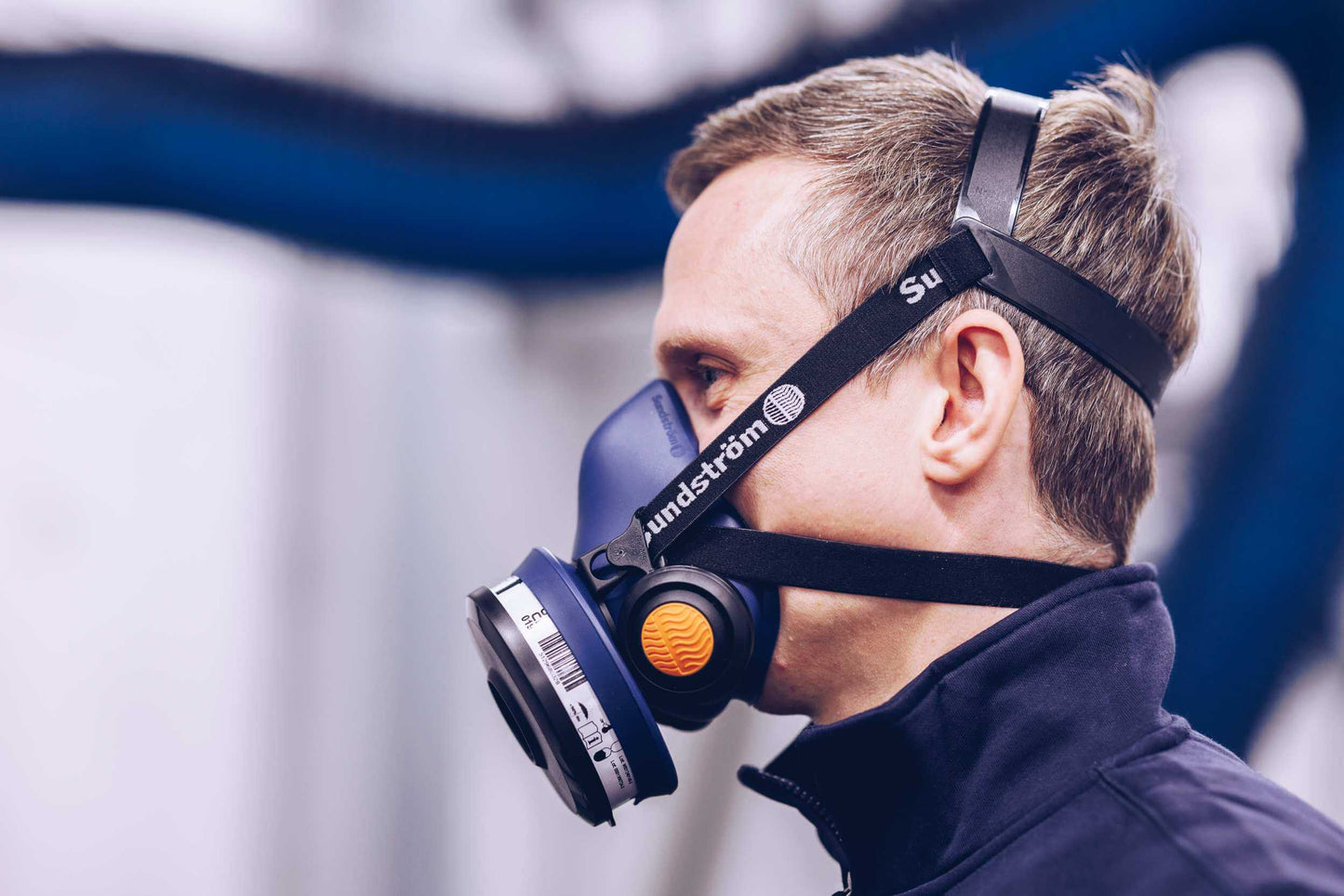 A side profile of a man wearing a blue Sundström SR 100 half-mask respirator with a black head harness strap.