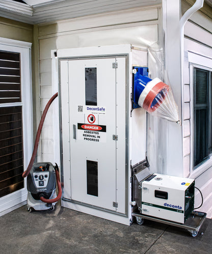 A DeconSafe decontamination unit set up against a building wall, featuring a "DANGER ASBESTOS REMOVAL IN PROGRESS" sign, a Nilfisk vacuum, and a portable deconta G55 air filtration unit.